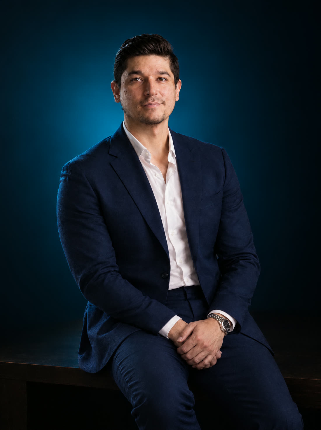 Dr. Alejandro Parra con traje azul marino y camisa blanca, sentado en un escritorio, sobre fondo azul